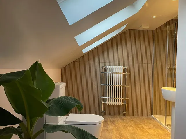 Loft bathroom under two roof windows, with wood slat wall panelling, a traditional column radiator, a back-to-wall toilet and a large banana plant in the foreground