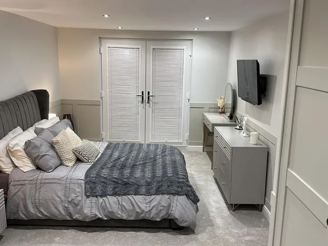 Bedroom seen between open wardrobe doors, with a grey velvet bed, double doors with louvred shutters, a dressing table and a wall-mounted TV