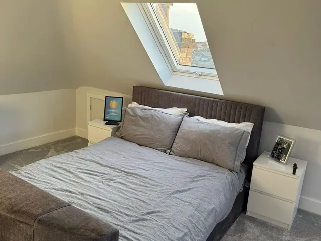 Loft bedroom with a grey upholstered bed beneath a roof window looking out over neighbouring rooftops