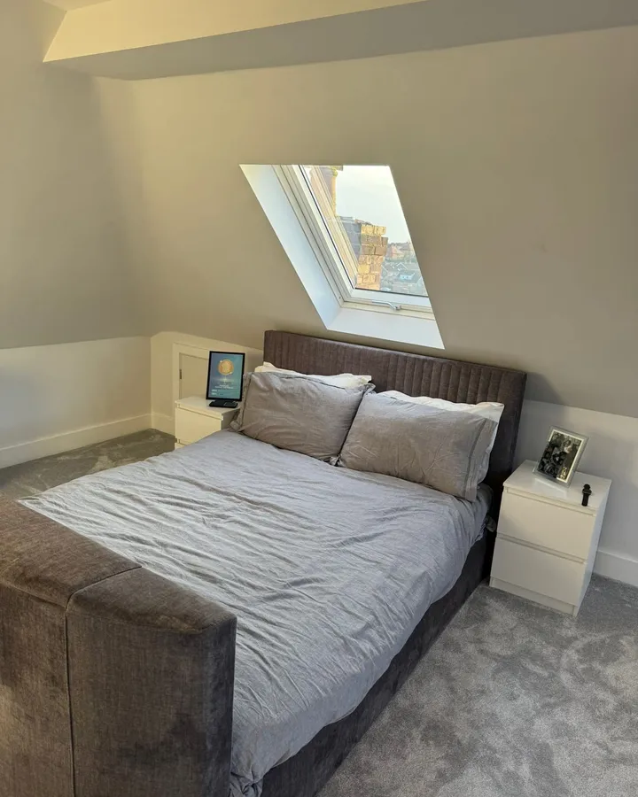 Loft bedroom with a grey upholstered bed beneath a roof window looking out over neighbouring rooftops