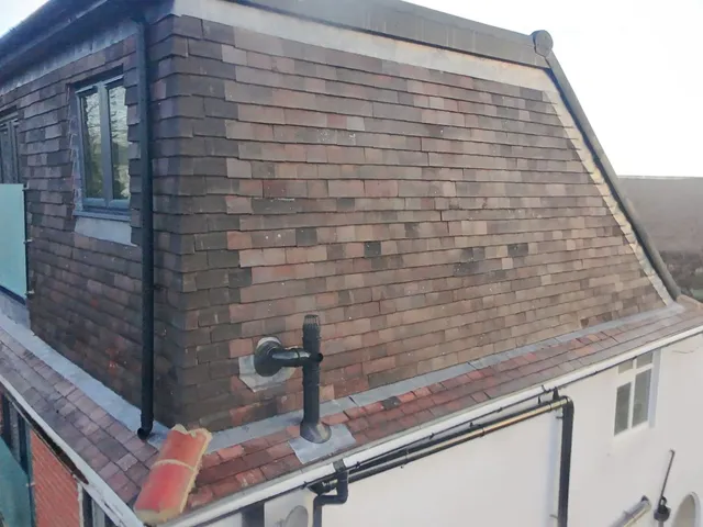 Close aerial view of the side of a dormer hung with multi-tone clay tiles, with a flue outlet and a glimpse of the glass balustrade on the front