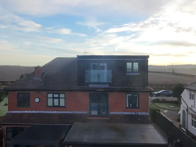 Aerial rear view of a red-brick semi-detached house with a tile-hung rear dormer and glass Juliet balcony above a flat-roofed single-storey extension, with fields beyond
