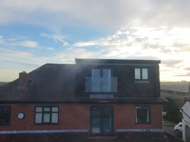 Red-brick house with a rear dormer clad in dark tiles and a glass Juliet balcony, under a hazy evening sky