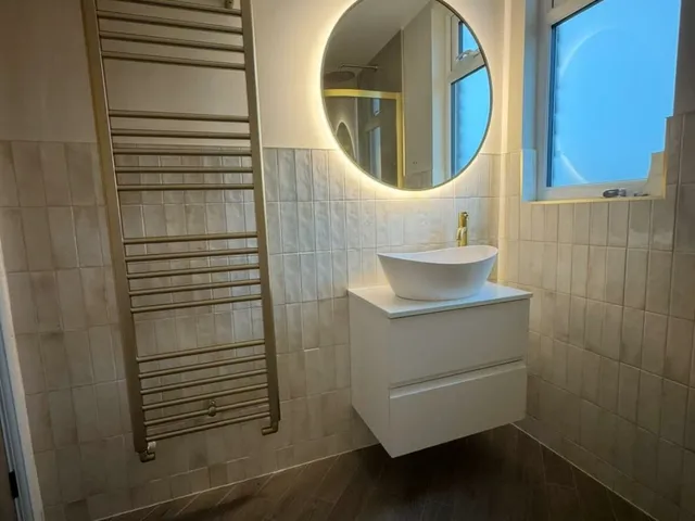Shower room with a round LED mirror, tall brass towel radiator and a floating white vanity with a vessel basin