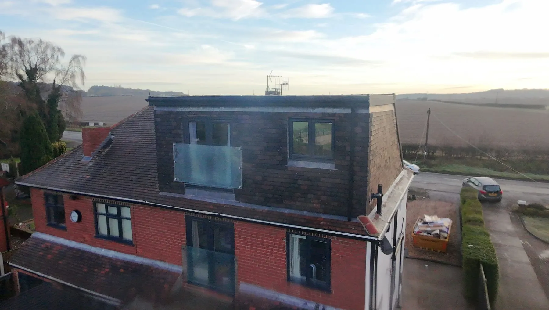 Flat-roofed dormer clad in dark hanging tiles with a glass Juliet balcony, above a red-brick house
